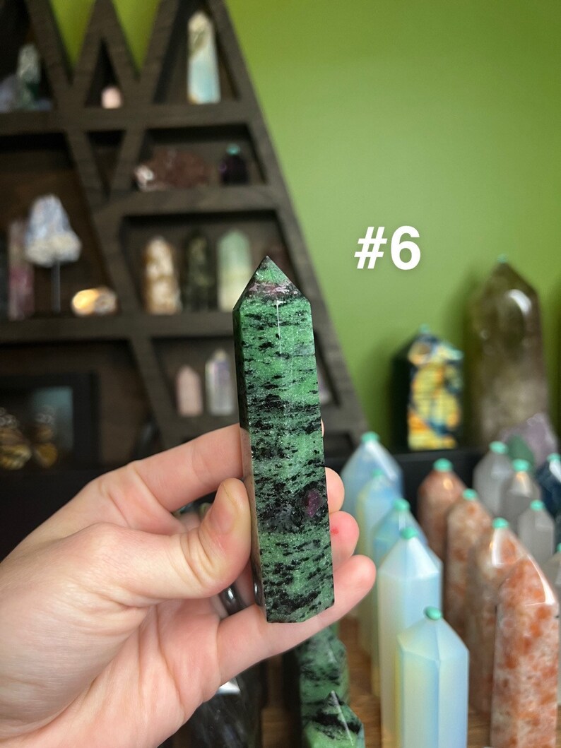 May include: A green and black zultanite point with a pink hue. The point is about 2 inches long and is held in a hand. The point is labeled #6.