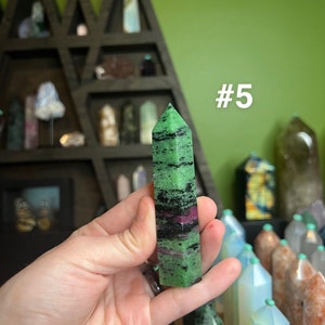 May include: A green, black, and pink gemstone point, also known as a crystal, is held in a hand. The point is about 2 inches long and has a pointed tip. The background is a shelf with other crystals and a green wall. The text #5 is visible in the image.