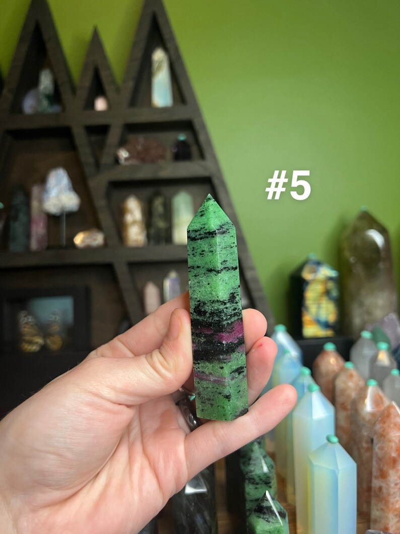 May include: A green, black, and pink gemstone point, also known as a crystal, is held in a hand. The point is about 2 inches long and has a pointed tip. The background is a shelf with other crystals and a green wall. The text #5 is visible in the image.