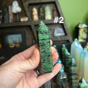 May include: A green and black gemstone point, labeled #2, is held in a hand. The point is a dark green color with black streaks throughout. The point is about 2 inches long.