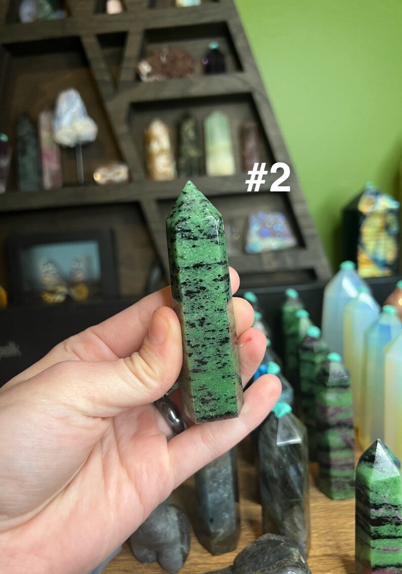 May include: A green and black gemstone point, labeled #2, is held in a hand. The point is a dark green color with black streaks throughout. The point is about 2 inches long.