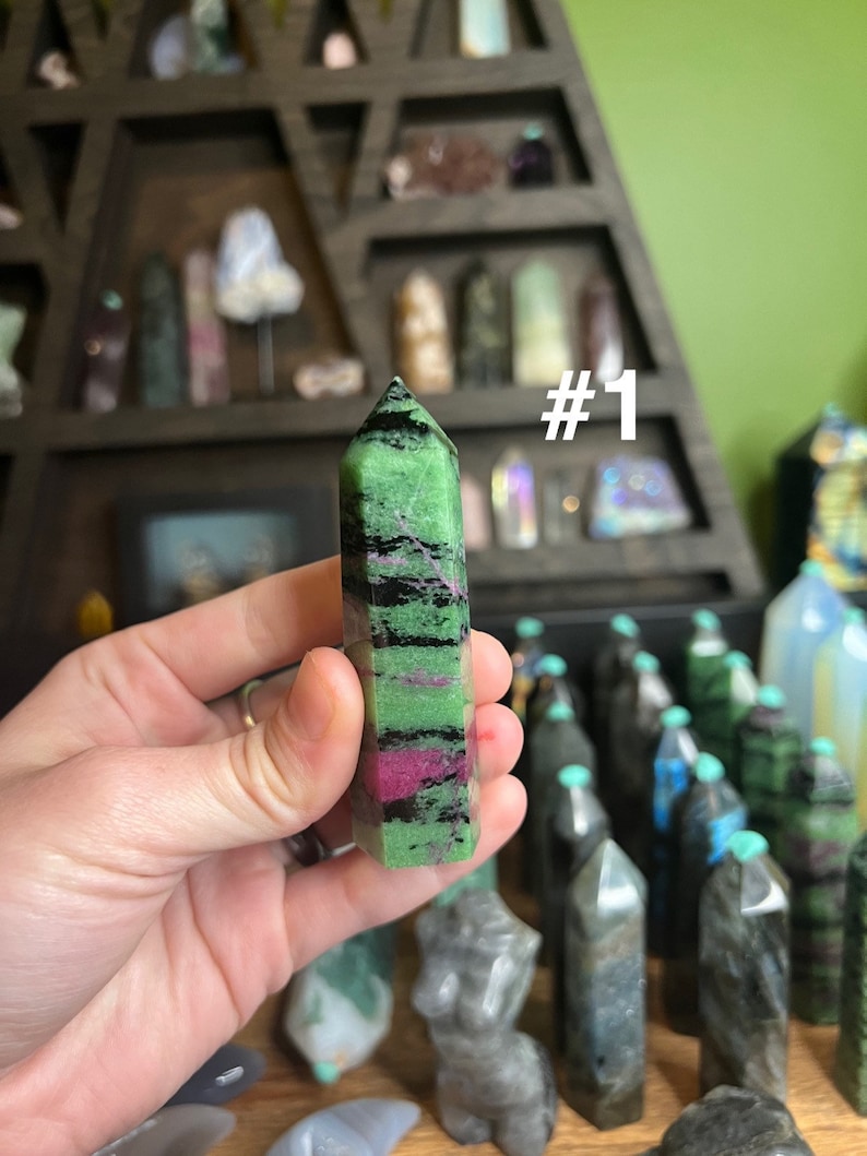 May include: A green, black, and pink crystal point with a #1 label. The crystal is being held in a hand. The crystal is surrounded by other crystals in a display case.