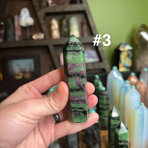 May include: A green, black, and pink crystal point with a pointed tip. The crystal is being held in a hand. The crystal is labeled #3.