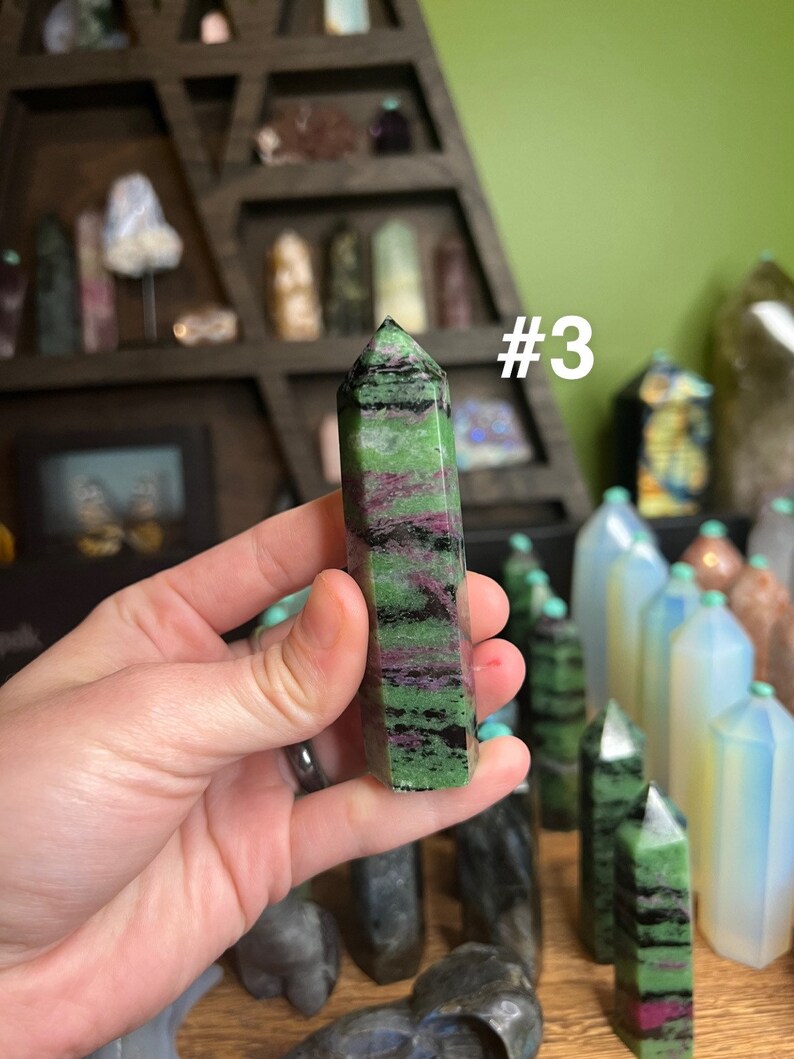 May include: A green, black, and pink crystal point with a pointed tip. The crystal is being held in a hand. The crystal is labeled #3.
