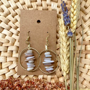 May include: A pair of gold hoop earrings with a stack of beige and white stones hanging from each hoop.