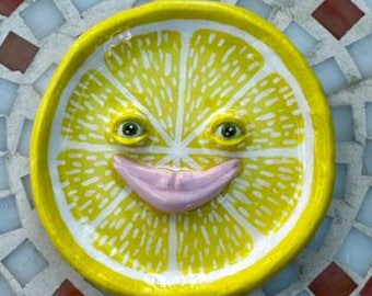 Könnte beinhalten: Eine skurrile, gelbe Keramik-Zitronenscheibe mit einem lächelnden Gesicht, vor einem Mosaikfliesen-Hintergrund. Die Zitrone hat grüne Augen und einen rosa Mund. Das Mosaik hat rote, weiße und blaue Fliesen.