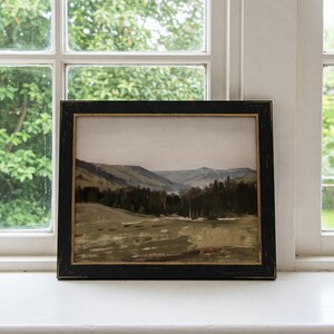 May include: A framed landscape painting depicting a valley with rolling hills and a forest in the distance. The painting is in a black frame with a gold trim.