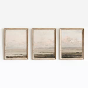 May include: A set of three framed landscape paintings with a muted color palette. The paintings depict a seascape with a cloudy sky and a distant shoreline. The frames are a light wood color.