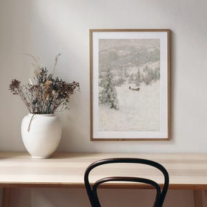 May include: A framed landscape art print depicts a snowy mountain scene with evergreen trees. The artwork is displayed on a light-colored wall above a wooden desk. A white vase with dried flowers sits on the desk.