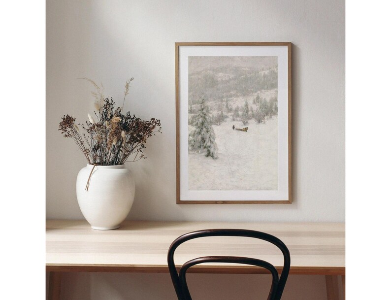 May include: A framed landscape art print depicts a snowy mountain scene with evergreen trees. The artwork is displayed on a light-colored wall above a wooden desk. A white vase with dried flowers sits on the desk.