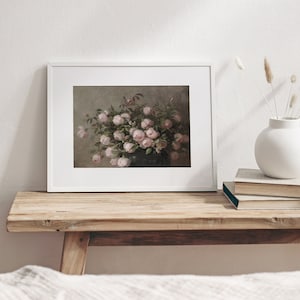 May include: A framed print featuring a still life of pink roses in a dark vase. The artwork is displayed in a white frame and rests on a rustic wooden bench. A white vase with dried flowers and a stack of books are also on the bench.