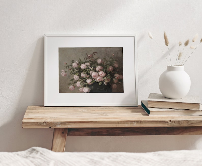 May include: A framed print featuring a still life of pink roses in a dark vase. The artwork is displayed in a white frame and rests on a rustic wooden bench. A white vase with dried flowers and a stack of books are also on the bench.