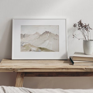 May include: A framed watercolor painting of a mountain range in shades of brown, beige, and green. The painting is on a wooden table with a vase of dried flowers and a stack of books.