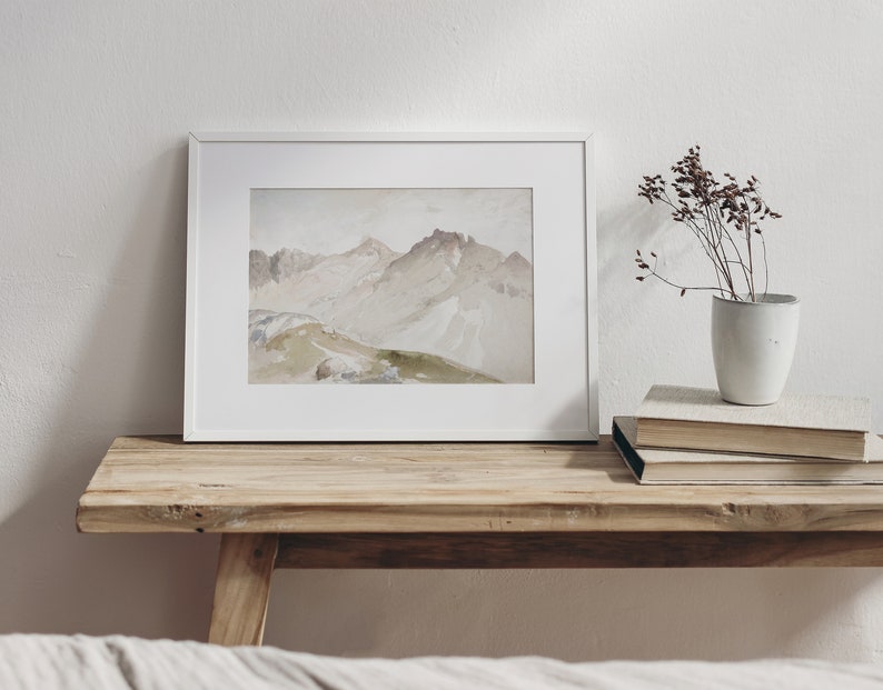 May include: A framed watercolor painting of a mountain range in shades of brown, beige, and green. The painting is on a wooden table with a vase of dried flowers and a stack of books.