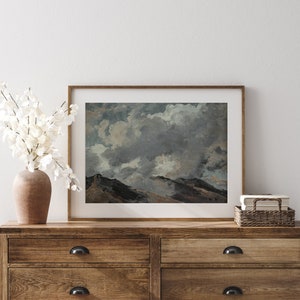 May include: A framed print of a landscape painting depicting a mountain range with a cloudy sky. The sky is a mixture of grey, white, and blue, with fluffy clouds. The mountains are a dark green and brown colour.