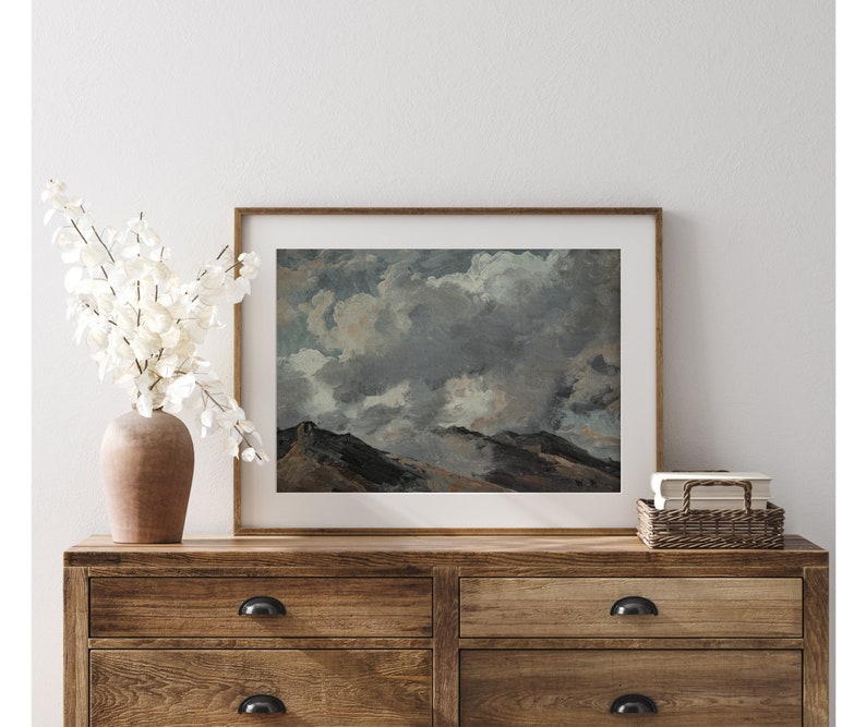 May include: A framed print of a landscape painting depicting a mountain range with a cloudy sky. The sky is a mixture of grey, white, and blue, with fluffy clouds. The mountains are a dark green and brown colour.