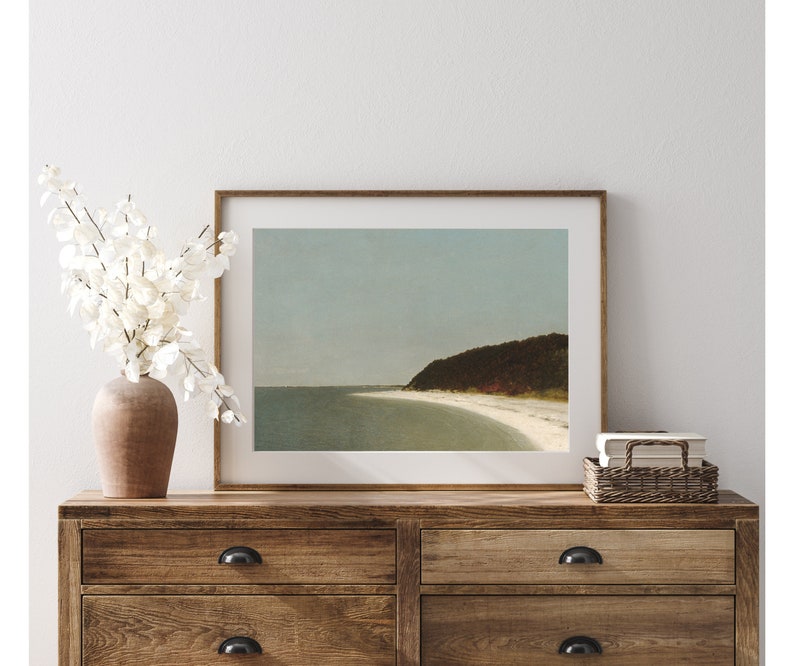May include: A framed print of a seascape with a sandy beach and a forested hill in the distance. The sky is a light blue and the water is a light turquoise.