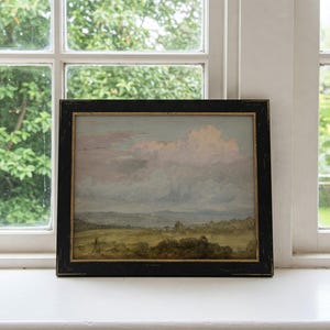 May include: A framed landscape painting depicting a field with a distant mountain range under a cloudy sky. The sky is a soft pink and blue, with fluffy white clouds. The field is green and brown, with a few trees scattered throughout.