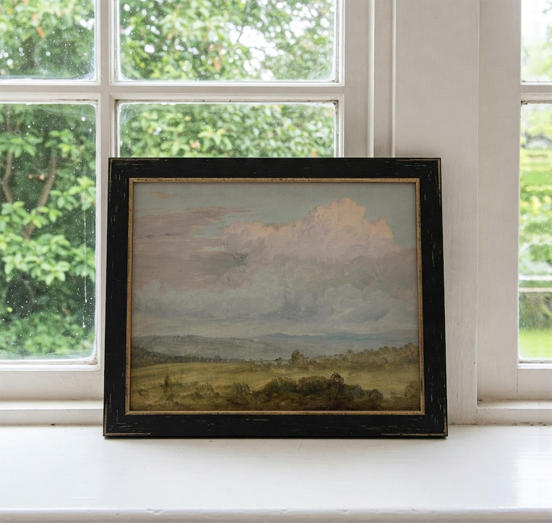 May include: A framed landscape painting depicting a field with a distant mountain range under a cloudy sky. The sky is a soft pink and blue, with fluffy white clouds. The field is green and brown, with a few trees scattered throughout.