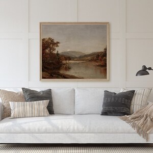 K&ouml;nnte beinhalten: Ein gerahmtes Landschaftsgem&auml;lde zeigt eine ruhige Flussszene mit herbstlichen B&auml;umen und fernen Bergen. Das Kunstwerk wird &uuml;ber einem wei&szlig;en Sofa mit dekorativen Kissen und einer neutralen Decke pr&auml;sentiert, wodurch eine gem&uuml;tliche Wohnzimmeratmosph&auml;re entsteht.