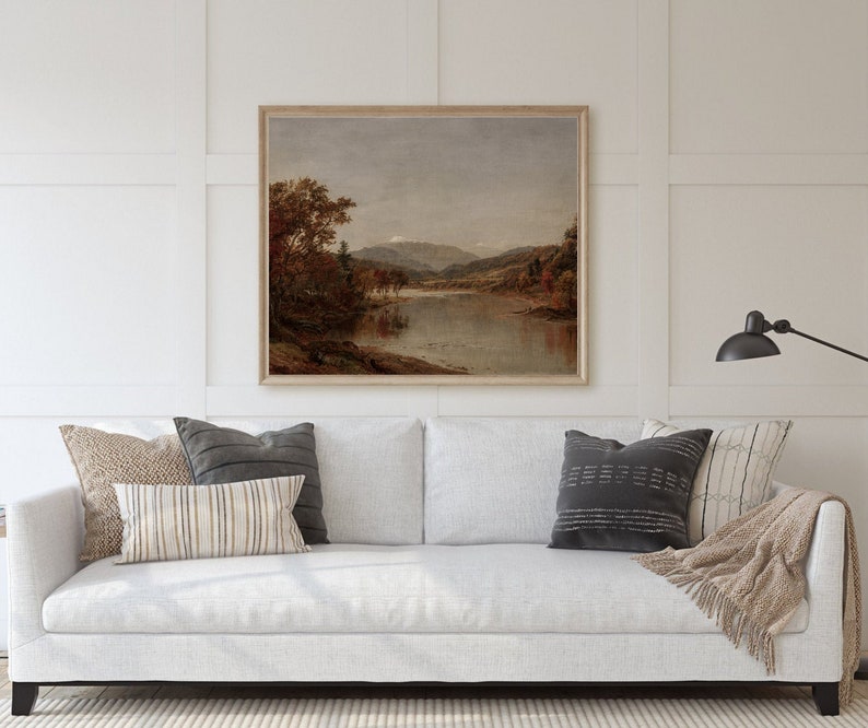 K&ouml;nnte beinhalten: Ein gerahmtes Landschaftsgem&auml;lde zeigt eine ruhige Flussszene mit herbstlichen B&auml;umen und fernen Bergen. Das Kunstwerk wird &uuml;ber einem wei&szlig;en Sofa mit dekorativen Kissen und einer neutralen Decke pr&auml;sentiert, wodurch eine gem&uuml;tliche Wohnzimmeratmosph&auml;re entsteht.