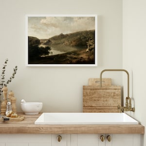May include: A framed landscape painting of a river winding through a wooded valley. The painting is in a white frame and is hanging on a light beige wall above a kitchen sink.