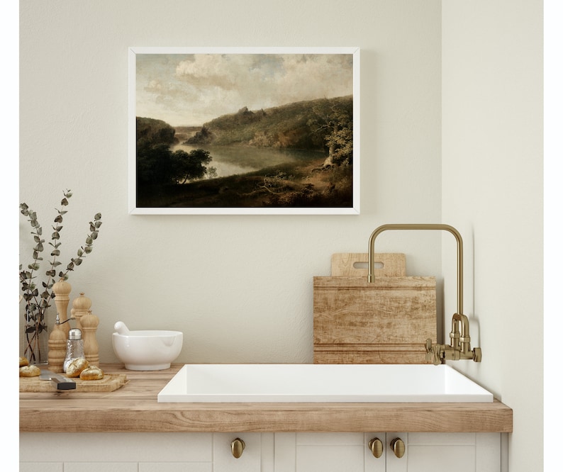 May include: A framed landscape painting of a river winding through a wooded valley. The painting is in a white frame and is hanging on a light beige wall above a kitchen sink.