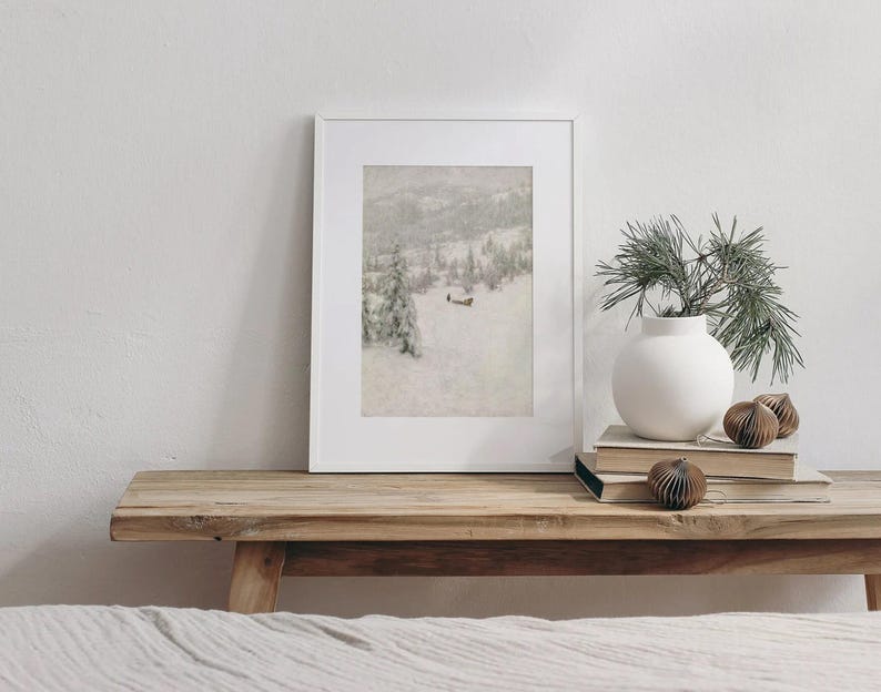 May include: A framed print of a snowy mountain landscape rests on a wooden bench. A white vase with greenery and decorative paper ornaments sit nearby. The scene evokes a sense of winter and nature.