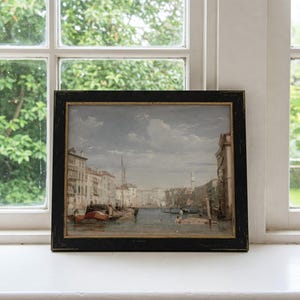 K&ouml;nnte beinhalten: Ein gerahmtes Aquarellbild, das eine Kanalszene in Venedig, Italien, darstellt. Das Bild zeigt einen schmalen Kanal mit Geb&auml;uden an beiden Seiten. Es befinden sich mehrere Boote im Wasser, darunter eine Gondel. Der Himmel ist hellblau mit einigen Wolken.