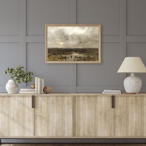 May include: A framed landscape painting depicting a field with a pond and birds flying overhead. The painting is in a wooden frame and is hanging on a gray wall above a wooden cabinet.