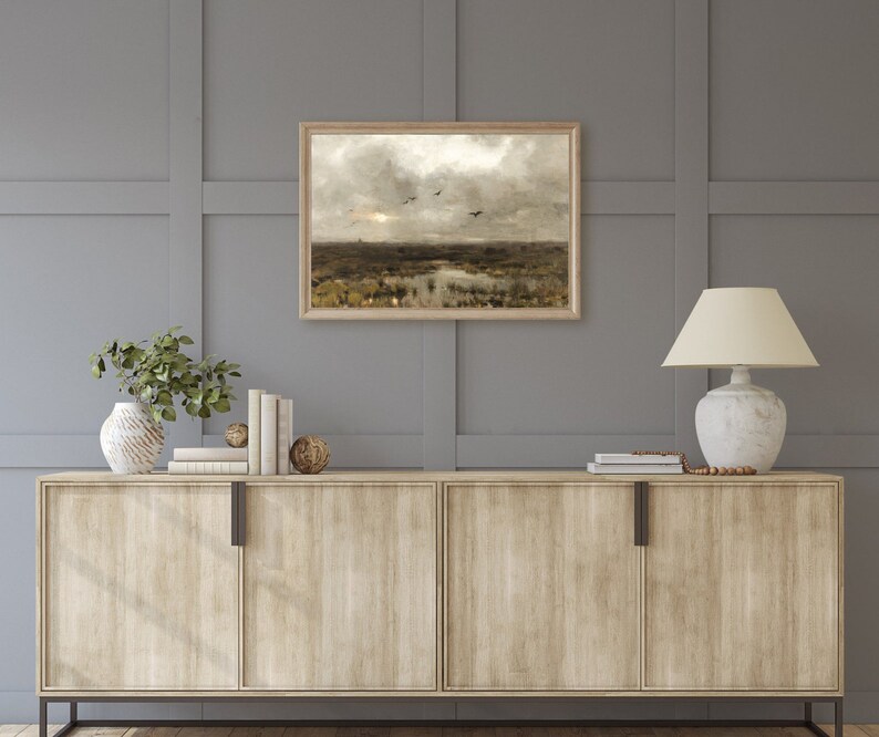 May include: A framed landscape painting depicting a field with a pond and birds flying overhead. The painting is in a wooden frame and is hanging on a gray wall above a wooden cabinet.