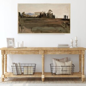 May include: A wooden console table with two shelves. The top shelf has a framed picture, candles, and a stack of books. The bottom shelf has two wire baskets, one with books and the other with pillows. The wall behind the table has a framed landscape painting.