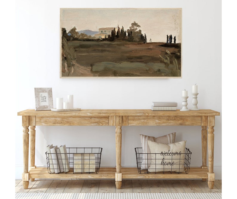 May include: A wooden console table with two shelves. The top shelf has a framed picture, candles, and a stack of books. The bottom shelf has two wire baskets, one with books and the other with pillows. The wall behind the table has a framed landscape painting.