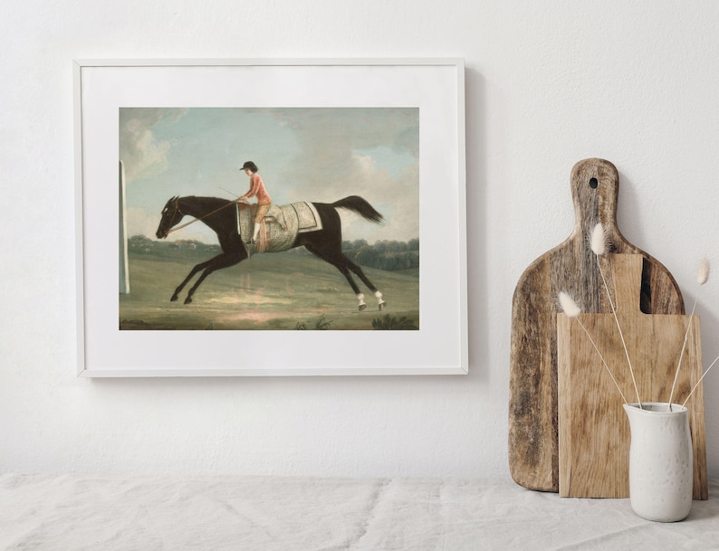 May include: Framed vintage equestrian art print. The artwork depicts a dark horse in motion with a jockey wearing a red jacket and black cap. The background features a green field and a cloudy sky.