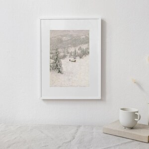May include: A framed print of a snowy landscape with a cabin and trees. The artwork is in a white frame and is displayed on a white wall. A white mug, a book, and a vase are on a table in front of the artwork.