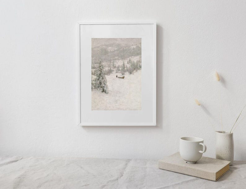 May include: A framed print of a snowy landscape with a cabin and trees. The artwork is in a white frame and is displayed on a white wall. A white mug, a book, and a vase are on a table in front of the artwork.