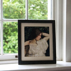 May include: A framed black and white portrait of a woman with long dark hair, wearing a white dress, looking down and resting her head on her arm.