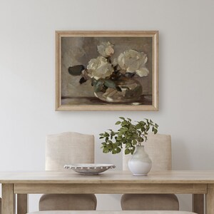 May include: A framed painting of white roses in a vase. The painting is in a light wood frame and is hanging on a white wall. The roses are in a clear glass vase with a white background. The painting is in a minimalist style.
