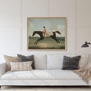 May include: A framed vintage equestrian painting depicts a jockey in orange atop a black horse mid-gallop. The artwork is displayed above a white sofa with decorative pillows and a throw blanket. A black lamp stands to the right.