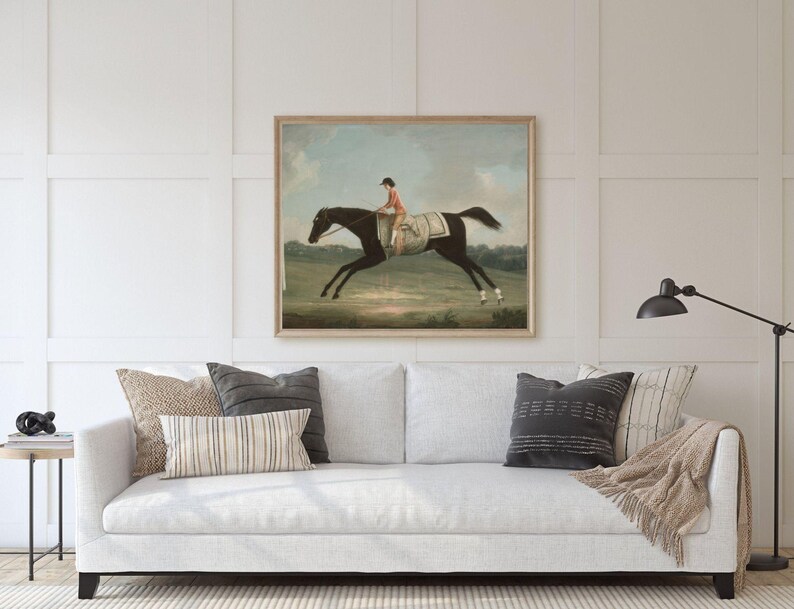 May include: A framed vintage equestrian painting depicts a jockey in orange atop a black horse mid-gallop. The artwork is displayed above a white sofa with decorative pillows and a throw blanket. A black lamp stands to the right.