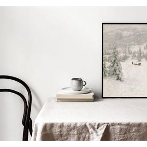 May include: A framed art print depicting a snowy landscape with trees and a small building. A gray mug and saucer sit atop a stack of books. A black chair is positioned to the left of the table, which is covered with a white tablecloth.