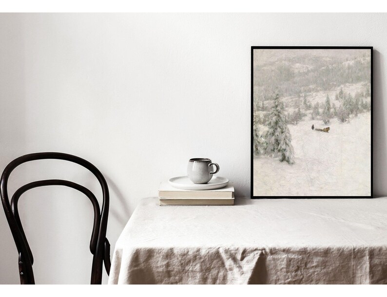 May include: A framed art print depicting a snowy landscape with trees and a small building. A gray mug and saucer sit atop a stack of books. A black chair is positioned to the left of the table, which is covered with a white tablecloth.