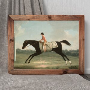 May include: A framed print of a horse race scene. The artwork depicts a jockey in a red jacket riding a black horse. The horse is leaping across a green field. The frame is made of wood.