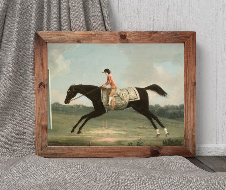May include: A framed print of a horse race scene. The artwork depicts a jockey in a red jacket riding a black horse. The horse is leaping across a green field. The frame is made of wood.