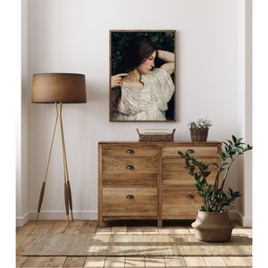 May include: A framed painting of a woman with long dark hair wearing a white dress, standing in a forest setting. The painting is hanging on a white wall above a wooden dresser with three drawers.