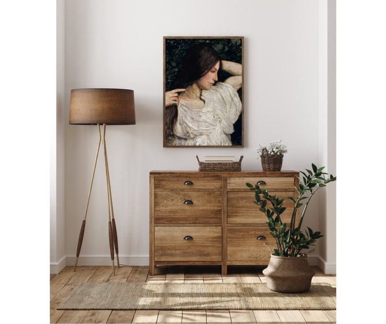 May include: A framed painting of a woman with long dark hair wearing a white dress, standing in a forest setting. The painting is hanging on a white wall above a wooden dresser with three drawers.