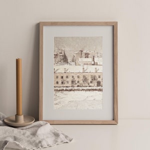 May include: A framed watercolor painting of a snowy cityscape with buildings and trees. The buildings are painted in shades of brown and white, and the trees are painted in shades of green and brown. The painting is set against a white background.