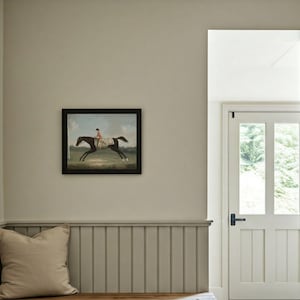 May include: A framed equestrian painting depicts a horse and rider in a field. The artwork is in a black frame and hangs on a light gray wall. A beige pillow rests on a wooden bench below the painting.