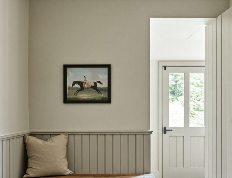 May include: A framed equestrian painting depicts a horse and rider in a field. The artwork is in a black frame and hangs on a light gray wall. A beige pillow rests on a wooden bench below the painting.