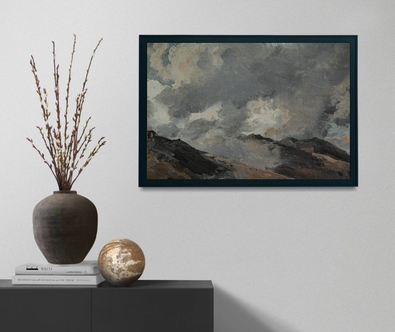 May include: A framed painting depicting a landscape with a cloudy sky and rolling hills. The sky is a mixture of grey and white, with glimpses of blue. The hills are a dark brown, with some areas of green and yellow. The painting is framed in a dark wood frame.